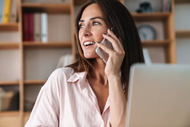 Ofiste dizüstü bilgisayarla çalışırken cep telefonuyla konuşan memnun bir iş kadını resmi