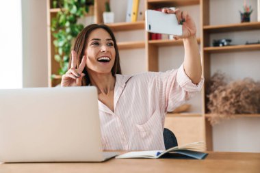 Ofiste dizüstü bilgisayarla çalışırken barış işareti yapan ve cep telefonundan selfie çeken mutlu yetişkin iş kadını görüntüsü
