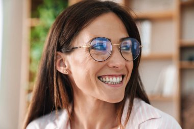 Ofiste poz verirken gülümseyen ve yan bakan gözlüklü neşeli iş kadınının yakın plan fotoğrafı.