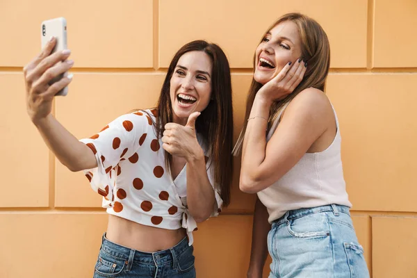 Neşeli yetişkin iki kadının cep telefonuyla selfie çekmesi ve şehirde poz verirken başparmağını kaldırması.