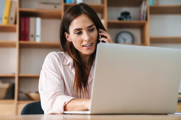 Ofiste Dizüstü Bilgisayarla Çalışırken Cep Telefonuyla Konuşan Memnun Bir Kadını — Stok fotoğraf