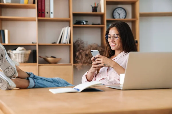 Image of smiling businesswoman using cellphone and laptop while sitting with legs on table in office