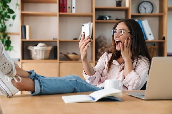 Image of excited woman screaming and taking selfie photo on cellphone while sitting with legs on table in office