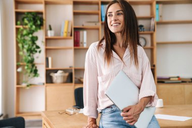 Ofiste otururken elinde dizüstü bilgisayarla gülümseyen neşeli bir iş kadını görüntüsü