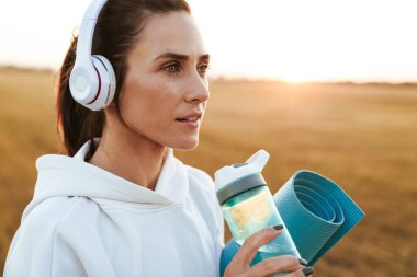 Kulaklık takmış, yoga minderini tutarken su içen güzel bir sporcunun görüntüsü.