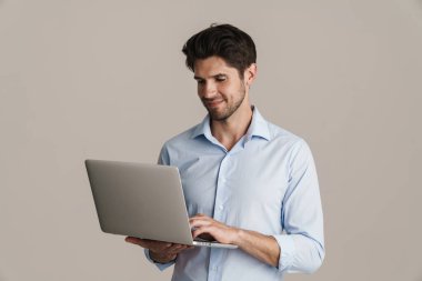 Sırıtkan esmer adamın bej arka planda dizüstü bilgisayarla çalışırken görüntüsü