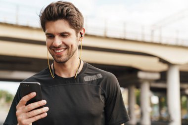 Kulaklıklı gülümseyen genç sporcunun şehir sokaklarında çalışırken cep telefonu kullanması.