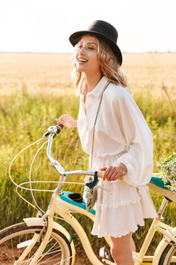 Kırsalda bisiklet sürerken gülümseyen ve elinde çiçek tutan genç sarışın kadın.
