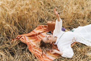 Genç bir kadının battaniyeye uzanmış, elini kaldırmış, piknik yaparken görüntüsü.