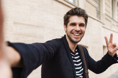 Genç ve yakışıklı bir iş adamının fotoğrafı. Sokakta yürürken kamerayla selfie çekiyor ve barış jesti gösteriyor.