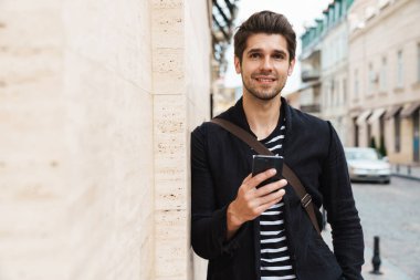 Şehir caddesinde duvara yaslanırken gülümseyen ve cep telefonu kullanan yakışıklı bir adam resmi