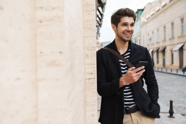 Şehir caddesinde duvara yaslanırken gülümseyen ve cep telefonu kullanan neşeli yakışıklı bir adam görüntüsü