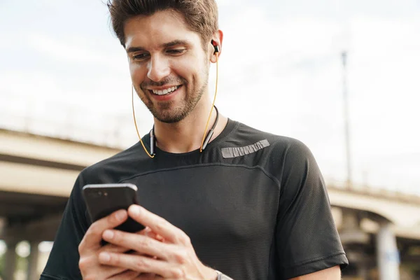 Obraz Uśmiechniętego Młodego Sportowca Słuchawkach Pomocą Telefonu Komórkowego Podczas Treningu — Zdjęcie stockowe