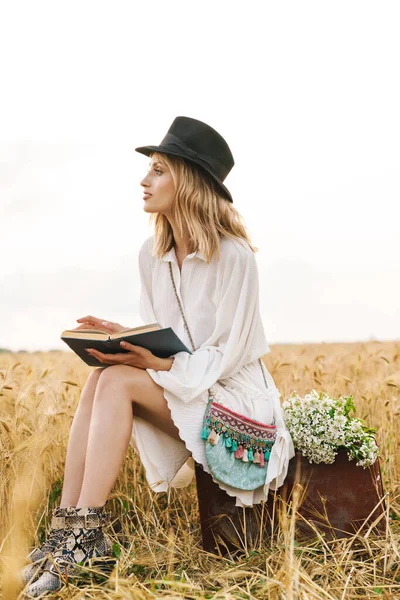 田舎の黄金のフィールドに座っている間若いブロンドの白人女性の読書本のイメージ — ストック写真