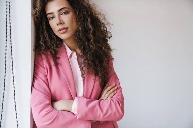 Beyaz odada duvara yaslanırken kameraya poz veren pembe ceketli güzel bir kadının fotoğrafı.