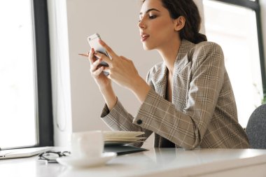 Ofiste dizüstü bilgisayarla çalışırken cep telefonu kullanan kendinden emin bir kadının fotoğrafı.