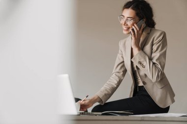 Ofiste dizüstü bilgisayarla çalışırken gözlüklü, gülümseyen esmer kadının cep telefonuyla konuşması.