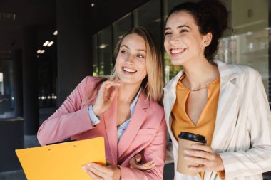 İki güzel, gülümseyen genç iş kadını şehir sokaklarında yürüyor, kahve içerken sohbet ediyorlar.