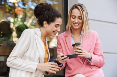 Dışarıda cep telefonu kullanan mutlu iki iş kadınının resmi.