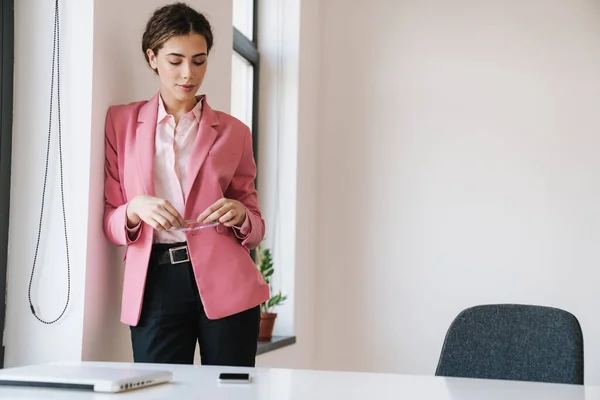 白いオフィスでカメラを構えながら眼鏡をかけながらピンクのジャケット姿の美しい女性の写真 — ストック写真