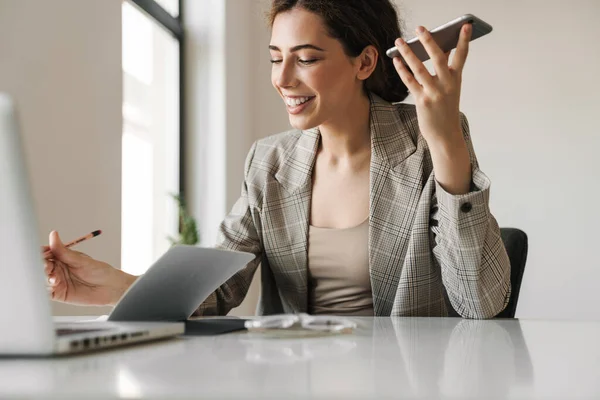 照片中快乐而友善的女人一边在办公室里与笔记本电脑一起工作一边笑着用手机 — 图库照片