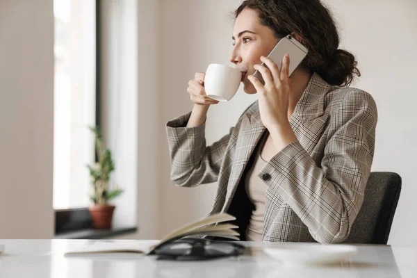 Photo Pleased Brunette Woman Drinking Coffee Talking Cellphone While Working — Stock Photo, Image