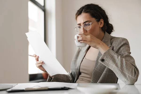 Foto Mujer Morena Enfocada Anteojos Bebiendo Café Mientras Trabaja Con — Foto de Stock