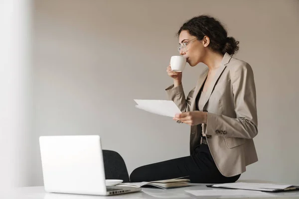 Photo of focused brunette woman in eyeglasses drinking coffee while working with laptop and documents at office