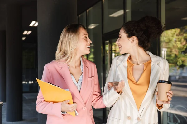 İki güzel, gülümseyen genç iş kadını şehir sokaklarında yürüyor, kahve içerken sohbet ediyorlar.