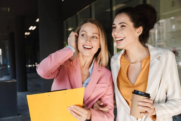 İki güzel, gülümseyen genç iş kadını şehir sokaklarında yürüyor, kahve içerken sohbet ediyorlar.