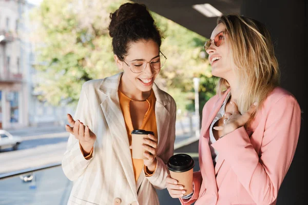 İki güzel gülümseyen genç iş kadını şehir sokaklarında yürüyor, kahve içerken konuşuyor, gülüyorlar.