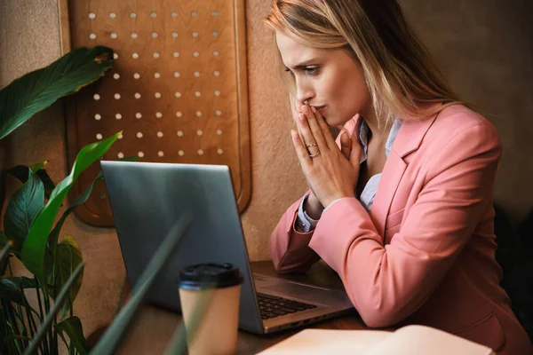 Attractive worried young businesswoman looking at laptop computer while sitting at the cafe table indoors