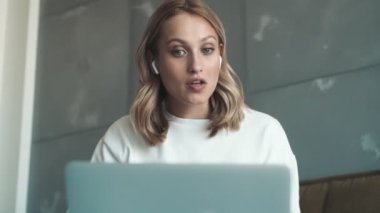 Kulaklıklı, gülümseyen genç bir kadın evinde oturan dizüstü bilgisayarını ve video bağlantısını kullanan biriyle konuşuyor.