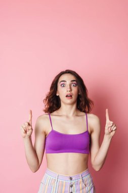 Sütyenli, pembe arka planda, parmaklarını yukarı gösteren kızıl saçlı bir kadın resmi.