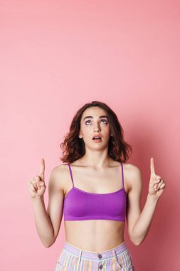 Sütyenli, pembe arka planda, parmaklarını yukarı gösteren kızıl saçlı bir kadın resmi.