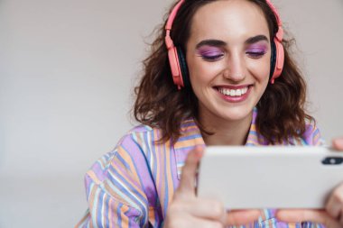 Beyaz arka planda izole edilmiş cep telefonu ve kablosuz kulaklık kullanan kızıl mutlu kadın görüntüsü