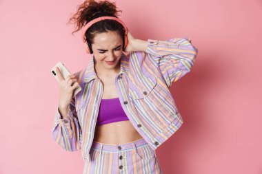 Pembe arka planda izole edilmiş cep telefonu ve kablosuz kulaklık kullanan kızıl mutlu kadın görüntüsü