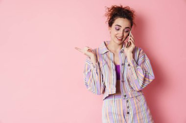 Cep telefonuyla konuşan ve pembe arka planda izole edilmiş parmağını gösteren kızıl saçlı bir kadın resmi.