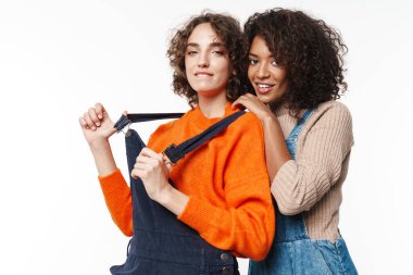 Güzel, mutlu, iyimser, kot tulumlu iki çok kültürlü kızın beyaz duvar arkasında izole edilmiş görüntüsü..