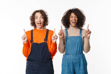 Beyaz duvar arkaplanından izole edilmiş kot elbiseli iki çok ırklı kız arkadaş telif uzayını işaret ediyor..