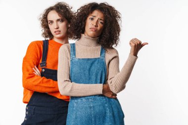 Kafası karışmış çok uluslu kadın portresi. Tulum giymiş, kameraya bakıyor ve beyaz arka planda izole edilmiş bir şekilde başparmağını işaret ediyor.