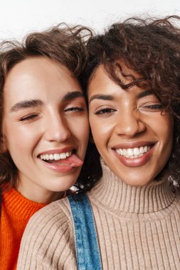 Güzel, mutlu, iyimser, kot tulumlu iki çok kültürlü kızın görüntüsü. Beyaz duvarın ardından izole edilmiş, dillerini gösteriyorlar..