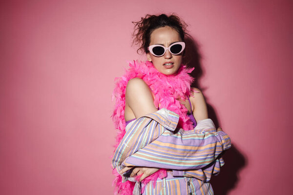 Image of caucasian displeased woman in sunglasses and boa looking at camera isolated over pink background