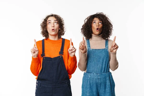 Beyaz duvar arkaplanı üzerinden izole edilmiş kot giysili iki çok ırklı kızın görüntüsü telif uzayını gösteriyor.