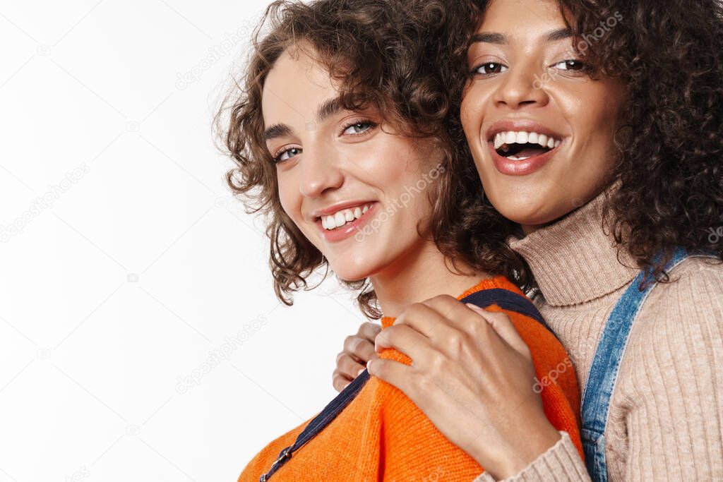 Portrait of cheerful multinational women in overalls laughing and hugging isolated over white background