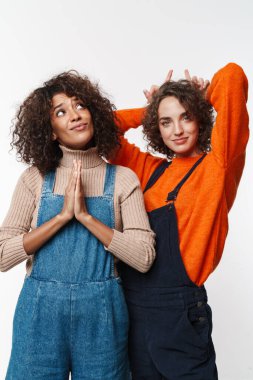 Güzel, mutlu, iyimser, kot tulumlu iki kız arkadaşın beyaz duvar arkasına izole edilmiş melek ve iblis hareketlerini gösteren görüntüsü..