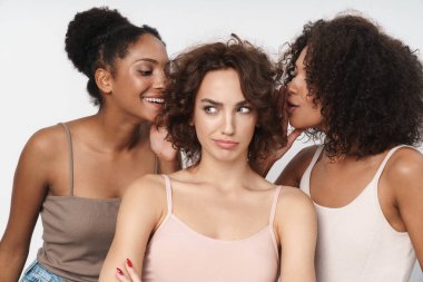 Üç etnik çeşitlilikte genç kadının portresi bir arada duruyor ve beyaz arka planda izole edilmiş bir şekilde kızların kulaklarına sırlar fısıldıyor.