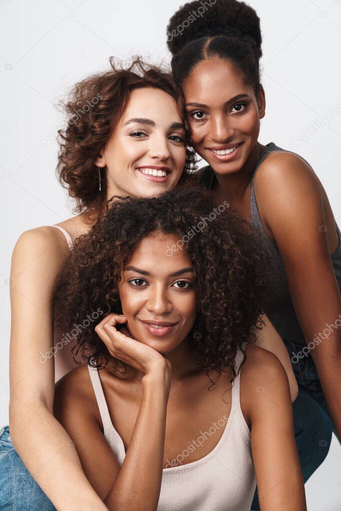 Portrait of three beautiful multiethnic women standing together and smiling at camera isolated over white background