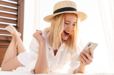 Hasır şapkalı heyecanlı bir sarışının, otelde yatakta birinci sınıf jest yaparken cep telefonu kullanması.