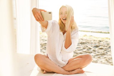 Yazın kumsalda oteldeyken cep telefonuyla selfie çeken neşeli sarışın bir kadın.